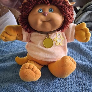 May include: A plush doll with a round tan face, blue eyes, and a dark brown yarn mane. It wears a light pink short-sleeved shirt featuring a small yellow bear patch and a brown collar with a round gold-colored pendant. The doll has orange limbs.