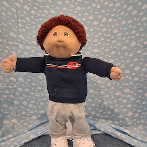 May include: A doll with brown yarn hair and a round face wearing a navy blue hooded sweatshirt with a red oval logo that reads athletics, grey sweatpants, and white shoes. The doll has a light skin tone, brown eyes, and a small dimple on the cheek.