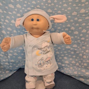 May include: A doll with a round face, blue eyes, and a light blue hat with pink-lined ears. It wears a light blue long-sleeved top with a snap closure, featuring a graphic of a character, a yellow moon, and the text Welcome Tiny Cuddler.