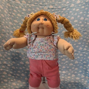 May include: A doll with blonde yarn hair styled in two braids with patterned ribbons. The doll has blue eyes, a round face, and dimples. It wears a short-sleeved white shirt with a small floral print in pink, purple, and green, paired with solid pink pants and light pink socks.