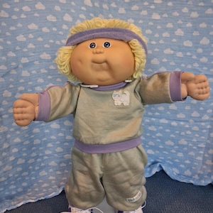 May include: A doll with blonde yarn hair wearing a purple headband, a light gray long-sleeved sweatshirt with a white cat graphic, matching gray pants with a purple waistband, and white sneakers with purple accents, standing with arms outstretched.