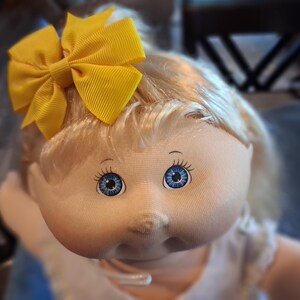 May include: A doll with light skin, blonde hair, and bright blue eyes. The doll features a soft fabric face and is wearing a large, bright yellow ribbon bow made of grosgrain fabric attached to the side of its hair.