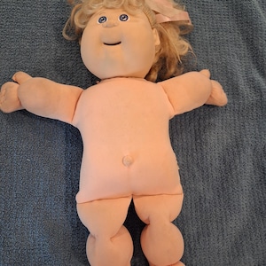 May include: A soft-bodied doll with peach-colored fabric skin, blonde yarn hair tied with a light pink ribbon, and blue eyes. The doll features a rounded face, a small nose, a stitched smile, and a prominent belly button on its torso.