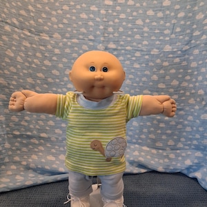 May include: A bald doll with a round face and blue eyes, wearing a light green and white striped short-sleeved shirt featuring a turtle graphic on the front, paired with light blue pants and white shoes, standing with arms outstretched.
