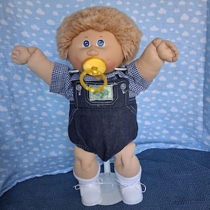 May include: A Cabbage Patch Kids doll with light brown yarn hair and blue eyes, wearing a blue and white gingham shirt under dark blue denim overalls. The doll has a yellow pacifier in its mouth, white socks, and white shoes, standing on a stand.
