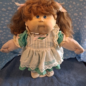May include: A soft-sculpture doll with curly auburn hair, blue eyes, and a round face. The doll wears a teal dress with puffy sleeves and a white pinafore featuring eyelet lace trim, light pink ribbons, and a signature embroidered on the hem.