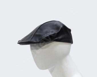 May include: A black leather flat cap featuring a structured crown and a short, stiff brim. The cap is crafted from a textured material with visible stitching along the seams, displaying a classic low-profile silhouette on a white mannequin head.