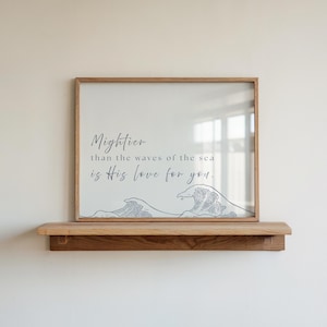 Puede incluir: Una impresión enmarcada con el texto "Más poderoso que las olas del mar es Su amor por ti". La impresión presenta un diseño de olas y se muestra en una estantería de madera. El marco es de color marrón claro.