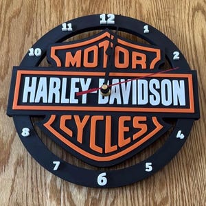 May include: Black and orange Harley-Davidson wall clock. The clock face features the Harley-Davidson logo with the words "MOTOR HARLEY-DAVIDSON CYCLES" in white and orange. The clock has white numbers and black hands.