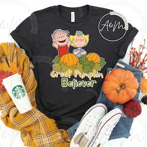 May include: Dark gray t-shirt featuring a cartoon graphic of two children standing behind pumpkins, with the text "Great Pumpkin Believer." The shirt is paired with jeans, white sneakers, and fall-themed accessories.