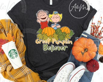 以下が含まれることがあります： 「Great Pumpkin Believer」の文字と、カボチャの後ろに立っている2人の子供の漫画のグラフィックが特徴の濃い灰色のTシャツ。ジーンズ、白いスニーカー、秋をテーマにしたアクセサリーが写っています。