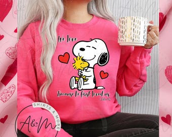 Könnte beinhalten: Ein pinkfarbener Pullover mit einer Grafik von Snoopy und Woodstock, die sich umarmen, mit dem Text "We Love Because He First Loved Us" und roten Herzen. Eine weiße Tasse mit goldenen Akzenten wird gehalten.