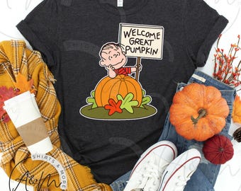 以下が含まれることがあります： 「Welcome Great Pumpkin」と書かれた看板を持った、カボチャの上に座っているキャラクターの漫画イラストが特徴の濃い灰色のTシャツ。デザインにはオレンジ、緑、赤のアクセントが含まれています。このシャツはジーンズと白いスニーカーと組み合わせています。