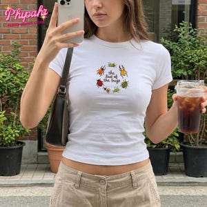 Può includere: Una t-shirt bianca a maniche corte con una grafica circolare di vari coleotteri colorati che circondano il testo "the last of the bugs" in un carattere semplice. La maglietta è abbinata a pantaloni color marrone chiaro e una borsa a tracolla marrone.