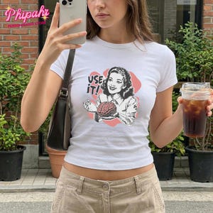 May include: White short-sleeved t-shirt featuring a vintage illustration of a woman holding a brain, with the text "USE IT!". The graphic is in black, red, and pink. The shirt is a fitted style.