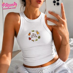 May include: A white ribbed tank top featuring a circular graphic of various colourful insects surrounding the text "the last of the bugs" printed in a dark brown font. The sleeveless garment has a high crew neckline and a fitted silhouette.