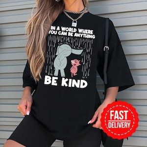 May include: Black t-shirt featuring the text "IN A WORLD WHERE YOU CAN BE ANYTHING BE KIND" and a graphic of an elephant and pig under a rain design. The shirt has a round neck and short sleeves.