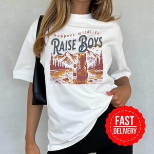 Peut inclure: T-shirt blanc avec un motif "Support Wildlife Raise Boys". Le motif représente un ours et un enfant debout dans l'eau, avec des montagnes et des arbres en arrière-plan. Un autocollant rouge "Fast Delivery" est dans le coin inférieur droit.
