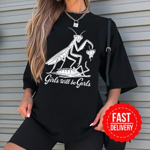 May include: Black t-shirt with a white graphic of praying mantises and the text "Girls will be Girls". The shirt has short sleeves and a loose fit. A red "Fast Delivery" sticker is also visible in the image.
