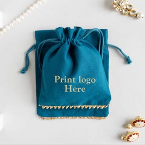 Könnte beinhalten: Türkisfarbener Kordelzugbeutel mit goldenem Besatz und dem Text "Print logo Here". Schmuck, darunter eine Perlenkette und Ohrringe, umgibt den Beutel auf einer weißen Oberfläche. Der Beutel ist aus weichem Stoff.