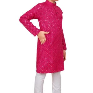 May include: A magenta kurta with a sequined pattern, paired with white trousers and brown loafers. The long-sleeved kurta has a mandarin collar and a pocket. The outfit is worn by a person wearing sunglasses.