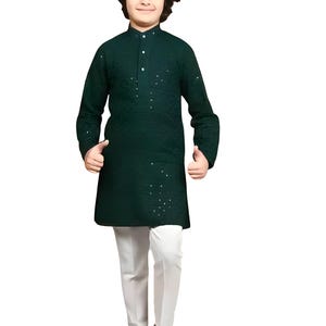 Op de afbeelding: Een jong persoon draagt een donkergroene tuniek met kleine, verspreide versieringen, gecombineerd met een witte broek en zwarte schoenen. De tuniek heeft een opstaande kraag en lange mouwen. De outfit is traditioneel.
