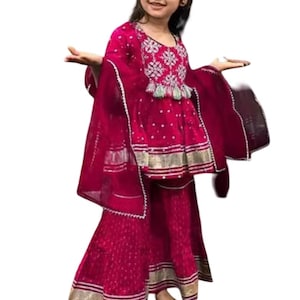 May include: A magenta-colored, traditional Indian outfit for a child. The ensemble includes a tunic with intricate embroidery, wide-legged pants, and a sheer scarf. Gold trim accents the edges of the outfit.