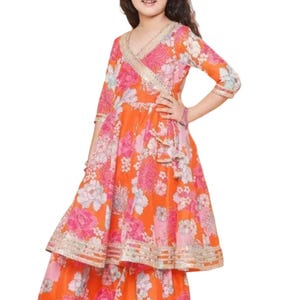 K&ouml;nnte beinhalten: Ein leuchtend orangefarbenes Kleid mit floralem Muster, Wickeloberteil und Stufenrock. Das Kleid hat rosa und wei&szlig;e Blumenmuster, goldfarbene Verzierungen und Dreiviertel&auml;rmel. Das Outfit ist f&uuml;r ein Kind entworfen.