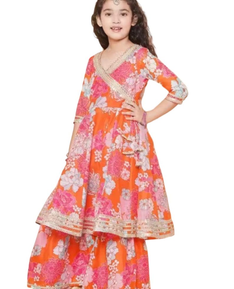 K&ouml;nnte beinhalten: Ein leuchtend orangefarbenes Kleid mit floralem Muster, Wickeloberteil und Stufenrock. Das Kleid hat rosa und wei&szlig;e Blumenmuster, goldfarbene Verzierungen und Dreiviertel&auml;rmel. Das Outfit ist f&uuml;r ein Kind entworfen.