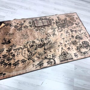 Puede incluir: Un mapa rectangular con apariencia de pergamino, con una detallada ilustraci&oacute;n en negro y marr&oacute;n de un mundo de fantas&iacute;a. El mapa incluye texto, un castillo, un lago y un barco. Es probable que el mapa sea un art&iacute;culo decorativo o un accesorio.