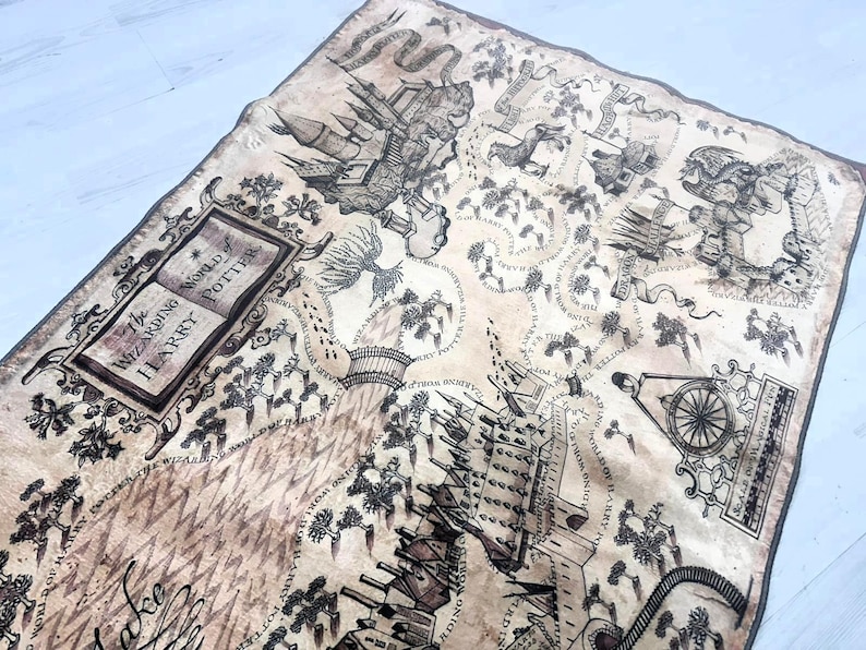 Puede incluir: Una manta rectangular de color marr&oacute;n claro con un dise&ntilde;o de mapa detallado, con ilustraciones y texto. El mapa incluye el texto "The Wizarding World of Harry Potter" e im&aacute;genes relacionadas. La manta tiene un borde marr&oacute;n oscuro.