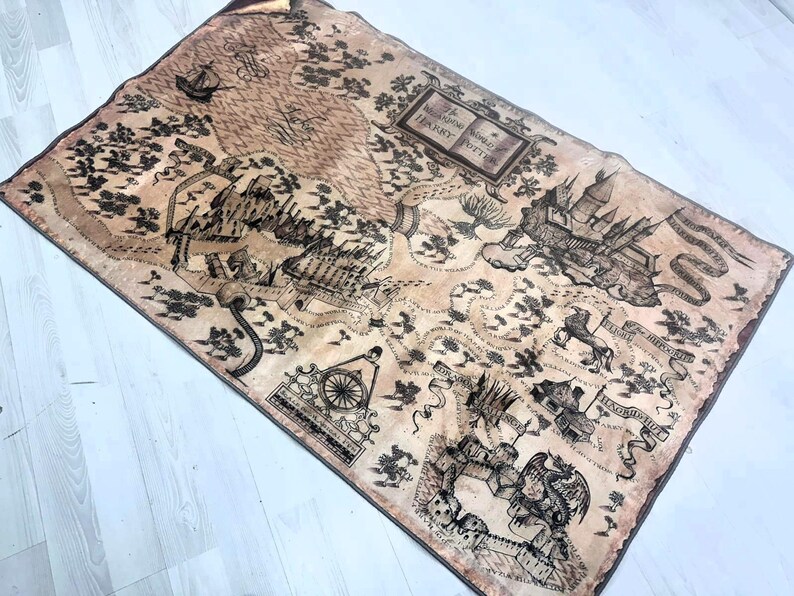 Puede incluir: Un mapa rectangular en tonos marr&oacute;n y beige, que representa un mundo de fantas&iacute;a con castillos, barcos y otras ilustraciones. El mapa incluye el texto "The Marauder's Map" y "Harry Potter". El mapa parece estar hecho de tela.