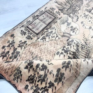 Puede incluir: Textil beige y negro con tem&aacute;tica de mapa con ilustraciones y texto de la serie Harry Potter. El mapa incluye el texto "The Wizarding World of Harry Potter" y un borde negro punteado.