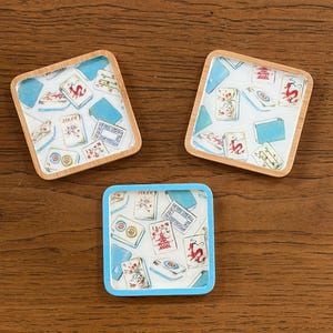 Puede incluir: Tres posavasos cuadrados con fondo blanco y un patrón repetitivo de fichas de mahjong, incluyendo dragones rojos, flores y fichas de comodín. Dos posavasos tienen marcos de madera clara y uno tiene un marco de color azul brillante.