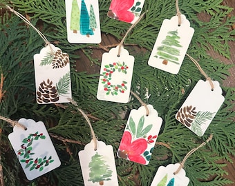 May include: A collection of white gift tags with watercolor designs, including Christmas trees, pine cones, and floral arrangements. Each tag is strung with twine and arranged on a bed of green foliage, perfect for holiday gifting.