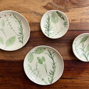 May include: Four round ceramic plates and bowls in varying sizes, featuring an off-white glaze with hand-painted green botanical illustrations of herbs like rosemary and sage. Each piece includes a series of small circular holes along the rim.