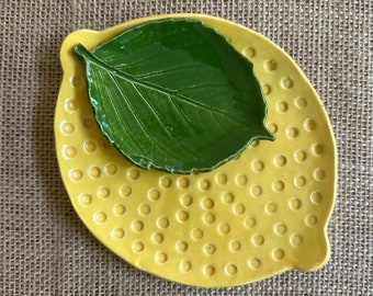 Op de afbeelding: Een gele keramische schotel in de vorm van een citroen met een groene schotel in de vorm van een blad erin. De grotere schotel heeft een getextureerd oppervlak met kleine cirkels.
