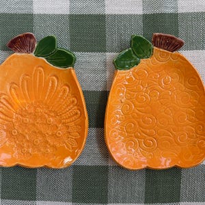 Puede incluir: Dos platos de cerámica naranja con forma de calabaza, cada uno con un tallo marrón y hojas verdes en la parte superior. Un plato tiene un patrón floral en relieve, mientras que el otro muestra un diseño texturizado de espirales.