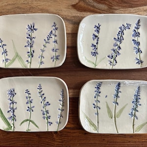 May include: Four rectangular ceramic plates with rounded corners, each featuring a hand-painted design of purple lavender sprigs with green stems and leaves on a cream-colored background. The plates have a smooth, glossy finish.