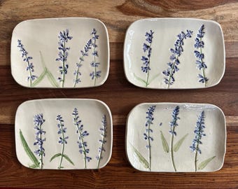 May include: Four rectangular ceramic plates with rounded corners, each featuring a hand-painted design of purple lavender sprigs with green stems and leaves on a cream-colored background. The plates have a smooth, glossy finish.