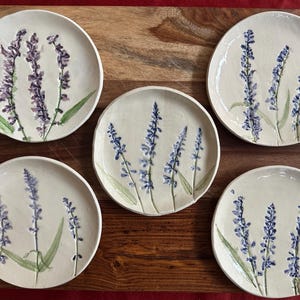 May include: Five ceramic plates with a cream-colored base, each adorned with a hand-painted lavender design. The lavender features purple flower spikes and green leaves. The plates are arranged on a wooden surface.