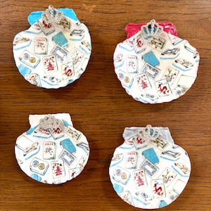 May include: Four scallop shells decorated with a white background featuring a pattern of colorful mahjong tiles, including a Joker tile. Each shell has a small, silver-toned crown embellishment attached to the top edge, painted in blue, red, or gray.