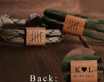 May include: Two bracelets with wooden accents. One bracelet features a camouflage-patterned cord, while the other has a green cord. The wooden accents are engraved with tally marks. The back of one accent shows initials and a date.