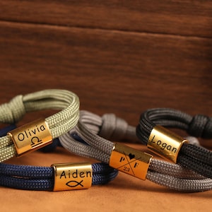 Può includere: Primo piano di diversi braccialetti in corda personalizzati. Ogni braccialetto presenta una chiusura in metallo color oro incisa con un nome e un simbolo. I braccialetti sono in vari colori, tra cui verde, blu, grigio e nero.