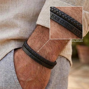 Può includere: Un bracciale in pelle nera con una fascia intrecciata e un segmento piatto liscio inciso con il nome Alexander. Un riquadro mostra un primo piano del bracciale con il testo YOUR CUSTOM TEXT inciso sulla superficie in pelle nera liscia.