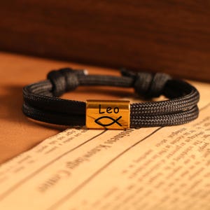 May include: Black rope bracelet with a gold-colored central bead. The bead is engraved with the word "Leo" and a fish symbol. The bracelet is resting on a vintage newspaper background.