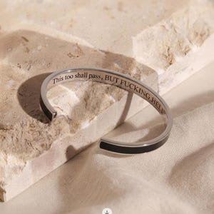 May include: A silver-toned metal cuff bracelet featuring a black inlay along the outer band. The interior surface is engraved with the black text "This too shall pass, BUT FUCKING HELL" in a serif font, visible along the inner curve of the bracelet.