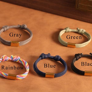 May include: Five bracelets in different colours: grey, green, rainbow, blue, and black. Each bracelet has a knotted design with a gold-coloured rectangular clasp. The words "Gray", "Green", "Rainbow", "Blue", and "Black" are printed on the image.
