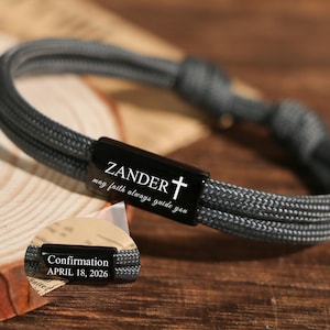 Puede incluir: Una pulsera trenzada gris con un cierre rectangular negro. El cierre presenta el nombre "ZANDER", una cruz y la frase "may faith always guide you". Una etiqueta más pequeña dice "Confirmation April 18, 2026".