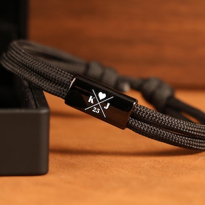 May include: Black rope bracelet with a black metal clasp. The clasp features the initials "K" and "J" with a heart symbol and the number "25" in white. The bracelet is displayed on a brown surface.